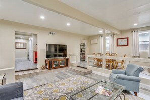 A spacious living room with a television, a glass coffee table, and a blue armchair. There is a dining area with wooden chairs and a table.