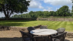 A round wooden table with wicker chairs in a garden with a stone wall and trees in the background. - Charlton Down Cottage - Westonbirt - Pet Friendly (Westonbirt)