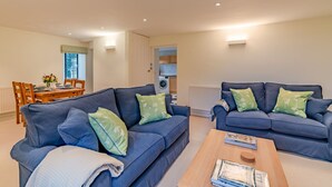 A modern living room with two blue sofas, green floral cushions, a wooden coffee table, and a dining area with a wooden table and chairs. - Charlton Down Cottage - Westonbirt - Pet Friendly (Westonbirt)