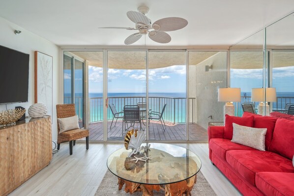 A modern living room with a glass coffee table, a red sofa, a ceiling fan, and a view of the ocean. - Hd1605 2 Bedroom Condo by RedAwning (Miramar Beach)
