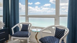 Una habitación con dos sillas blancas de mimbre, cojines azules estampados, una pequeña mesa redonda de vidrio y vistas al mar a través de amplios ventanales.