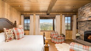 A wooden room with a bed, two plaid chairs, a fireplace, and a view of mountains.