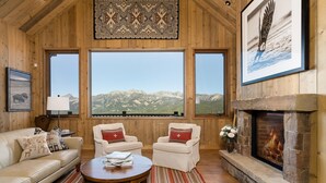 A cozy room with a fireplace, a sofa, two armchairs, a coffee table, and a large window offering a mountain view.