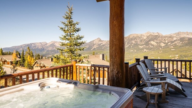 A hot tub on a wooden deck with a mountain view, wooden chairs, and a wooden railing.