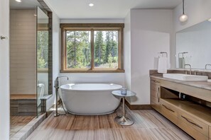 Een moderne badkamer met een vrijstaand bad, een douchecabine van glas, een groot raam met uitzicht op een bos, en een houten wastafel met een wastafel en spiegel.