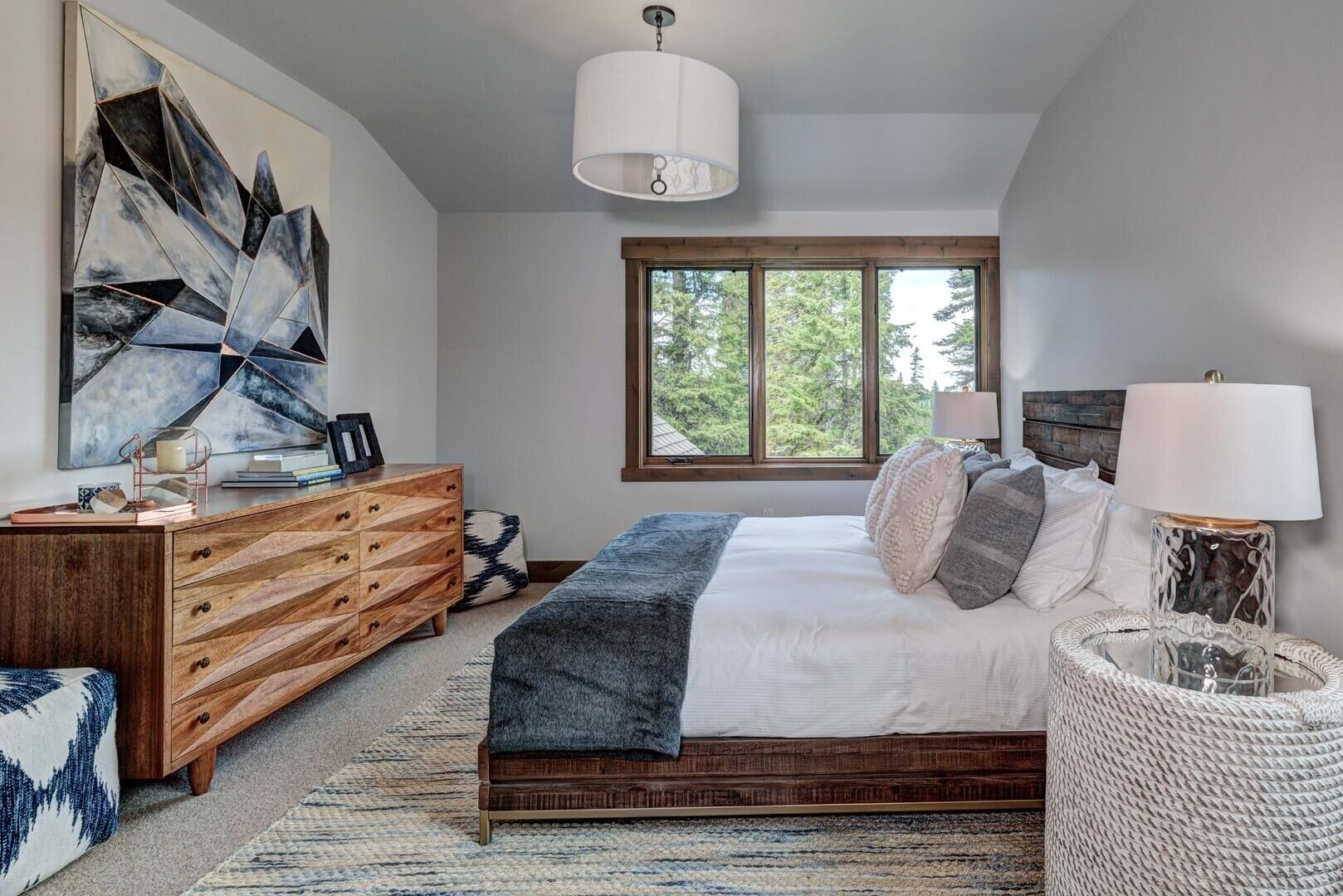A bedroom with a bed, wooden dresser, abstract artwork, a window with a view of trees, and a hanging light fixture.