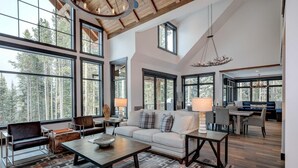 A spacious living area with a large window overlooking a forest, a cozy seating area, and a dining space. - 6 Jakes Lane 5 Bedroom Home by Moonlight Basin Lodging (Big Sky)