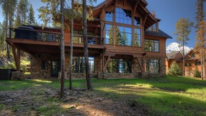 A multi-story log cabin with large windows and a stone foundation, surrounded by trees and a mountain backdrop. - 6 Jakes Lane 5 Bedroom Home by Moonlight Basin Lodging (Big Sky)