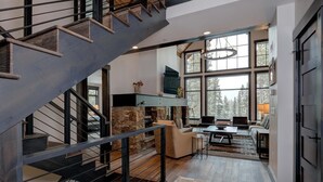 A modern interior with a staircase, stone fireplace, and large windows offering a view of trees. - 6 Jakes Lane 5 Bedroom Home by Moonlight Basin Lodging (Big Sky)