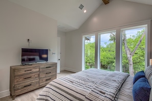 Un dormitorio con una cama grande, un televisor sobre un aparador de madera y vistas a árboles a través de ventanas amplias.