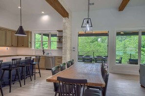 A modern kitchen with a large island, wooden bar stools, and a dining table with chairs. There are large windows offering a view of greenery.