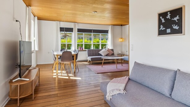 Ein modernes Wohnzimmer mit Holzboden, einem grauen Sofa, einem Esstisch mit Stühlen und einem TV-Schrank.