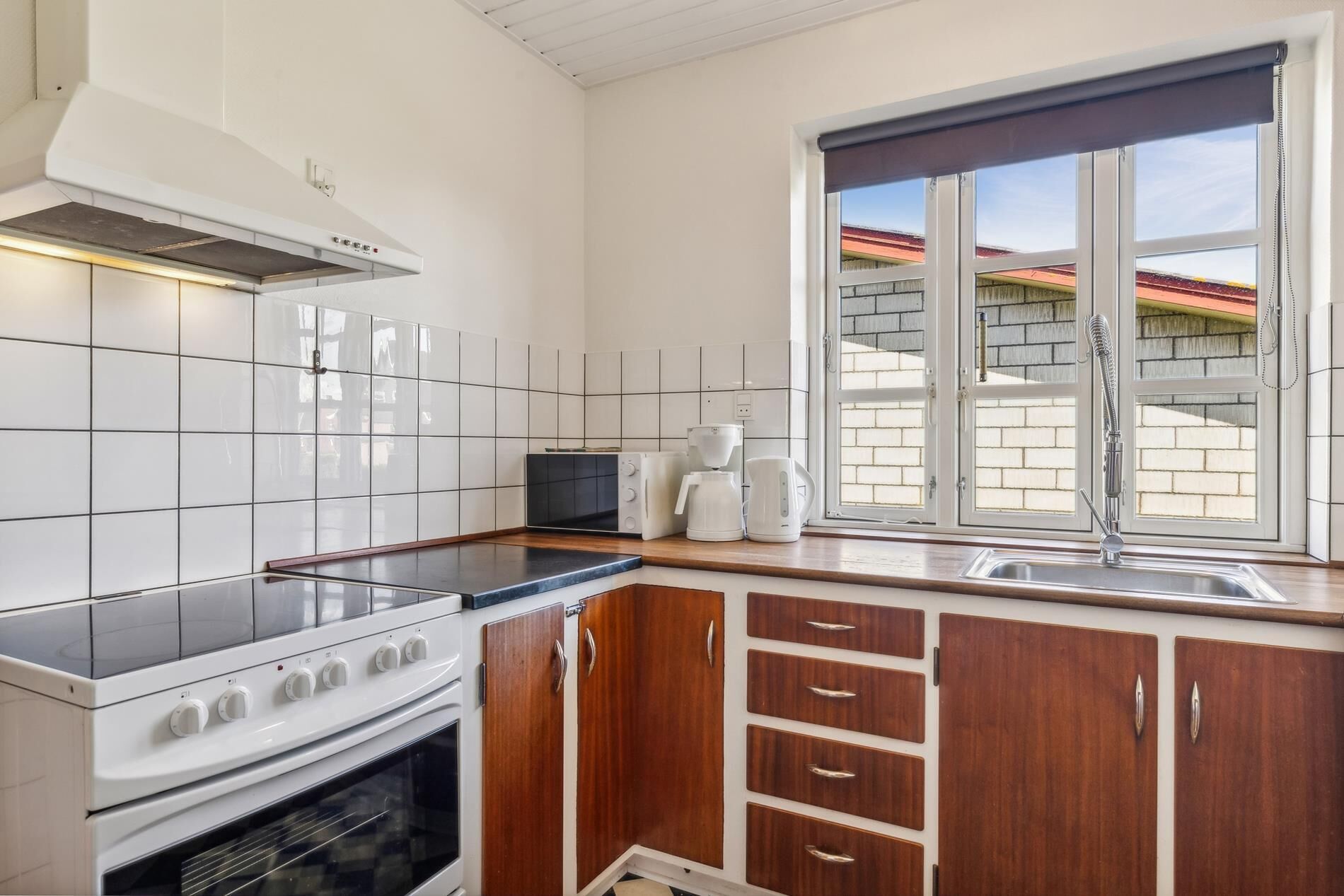 Una cocina con baldosas blancas, estufa, microondas, cafetera y ventana con vista a un edificio de ladrillo.