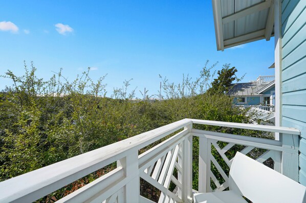 A white wooden deck with a railing overlooking a dense area of vegetation and a building with blue siding.