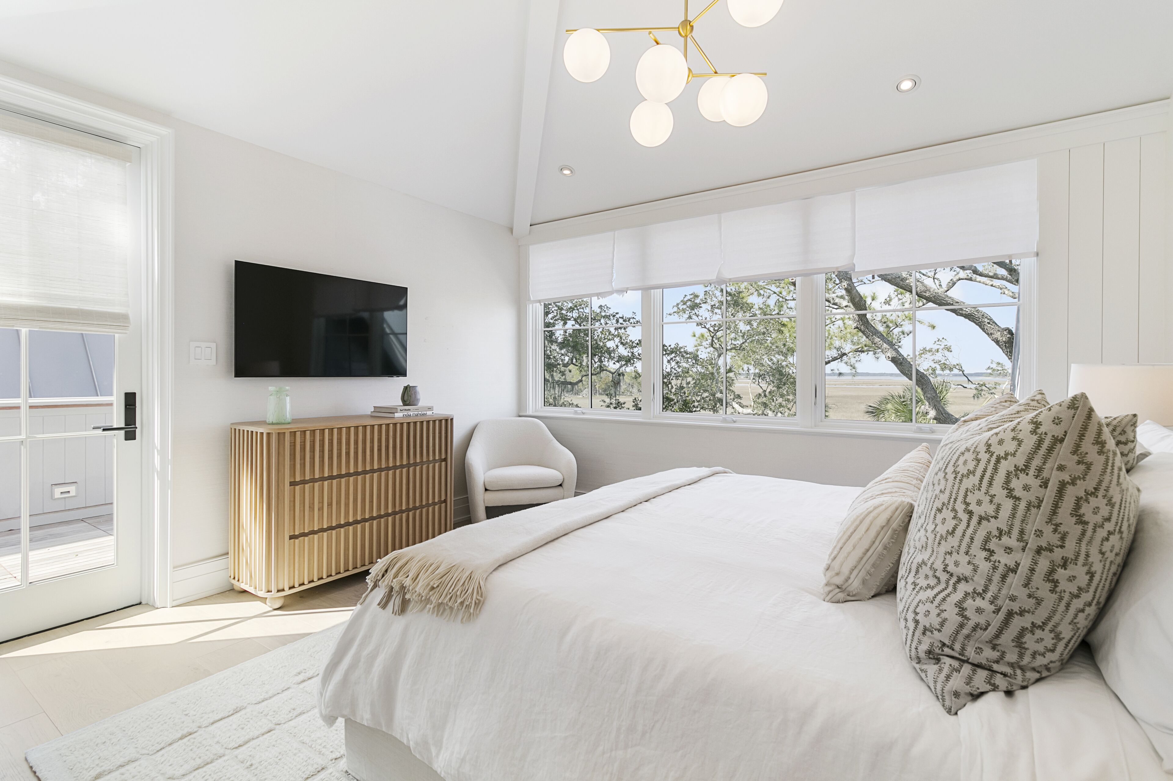 Un dormitorio con una cama grande, un televisor en la pared, un aparador de madera, una silla y un ventanal con vistas a árboles.