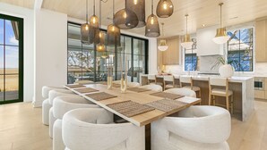 A modern dining area with a long wooden table, white upholstered chairs, and hanging pendant lights. - Personal Sanctuary, comfort and luxury at every turn (Kiawah Island)