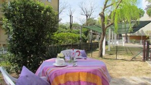 An outdoor dining area with a table set for two, surrounded by greenery and a building in the background. - Cozy condo 300mt from the beach (Bibione)