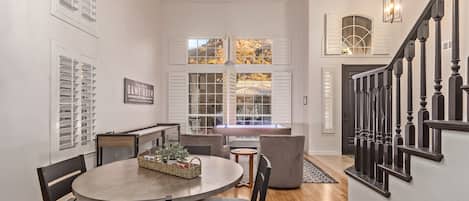 A dining area with a round table, chairs, and a basket with items. There are windows with shutters and a staircase with a railing.