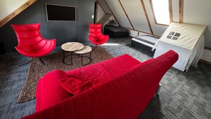 Un dormitorio tipo loft con techo inclinado, una cama con dosel blanco, dos sillas rojas, una mesita redonda pequeña y una alfombra gris.