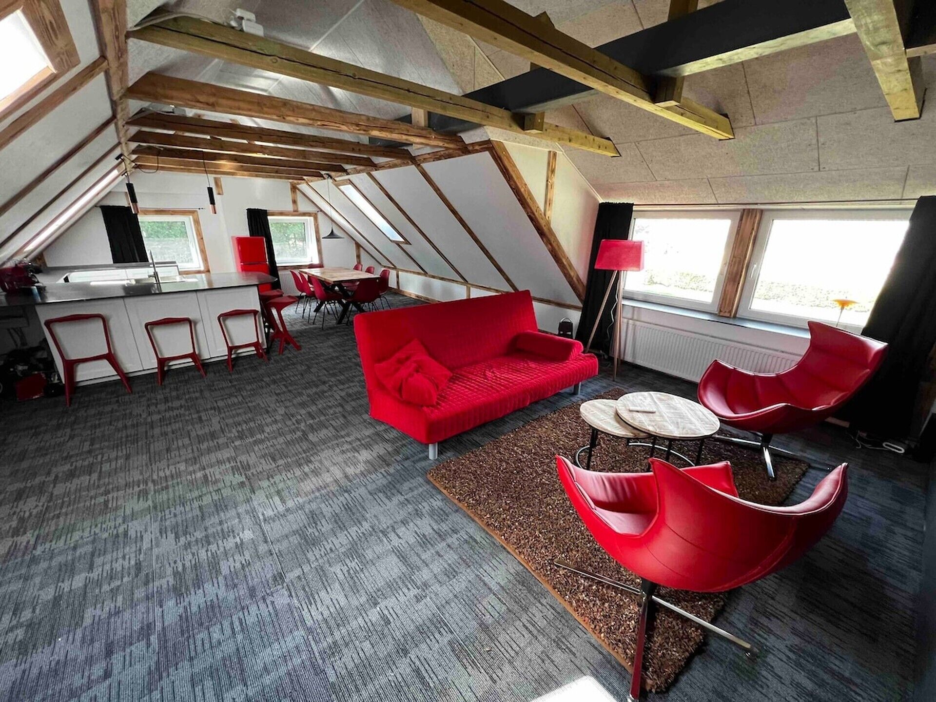 A modern loft-style room with a red sofa, red chairs, and a small round table. There is a kitchen area with a bar and stools, and a large window providing natural light.