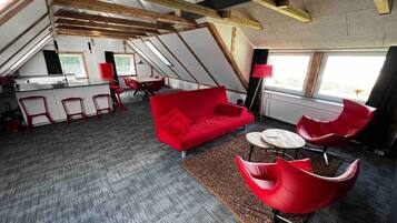 A modern loft-style room with a red sofa, red chairs, and a small round table. There is a kitchen area with a bar and stools, and a large window providing natural light.