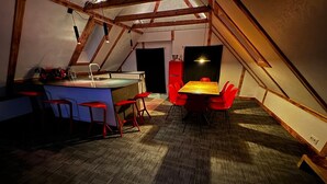 Una habitación acogedora en el ático, con techo de vigas de madera, un pequeño rincón de cocina con lavabo y taburetes, y una mesa de comedor con sillas rojas.