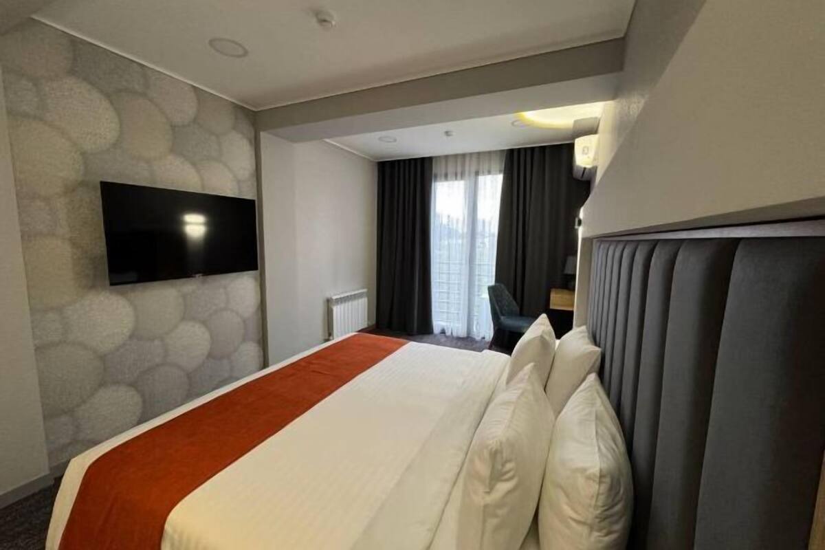 Habitación de hotel con cama, televisor de pantalla plana en la pared, ventana con cortinas y pared con diseño de piedra.