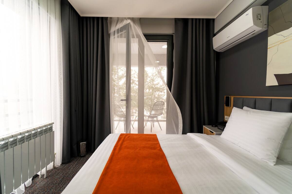 Una habitación de hotel con cama, mesita de noche, ventana con cortinas y un equipo de aire acondicionado.