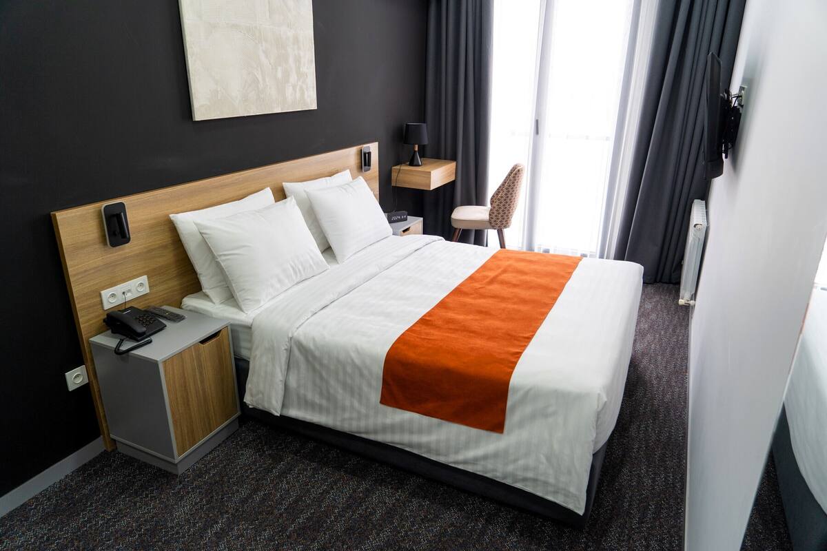 Habitación de hotel con cama, mesita de noche, lámpara, silla y vistas al exterior.