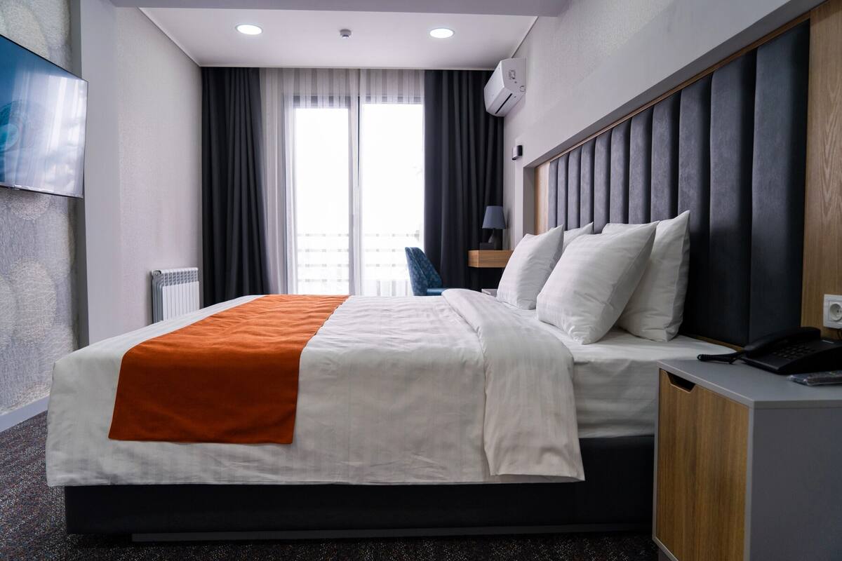 Habitación de hotel con una cama, una mesita de noche con un teléfono, una lámpara y vistas al exterior a través de una ventana.