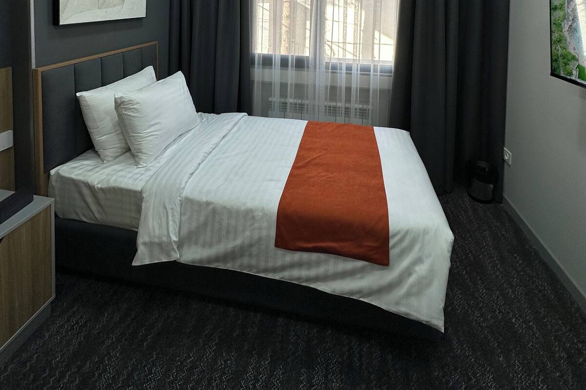 Habitación de hotel moderna con una cama bien hecha, un ventanal con cortinas translúcidas, un equipo de aire acondicionado y una obra de arte enmarcada en la pared.