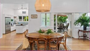 A spacious dining area with a wooden table and chairs, a hanging light fixture, and a view of the kitchen and outdoor patio. - The Grand Queenslander On Turner (Pimlico)