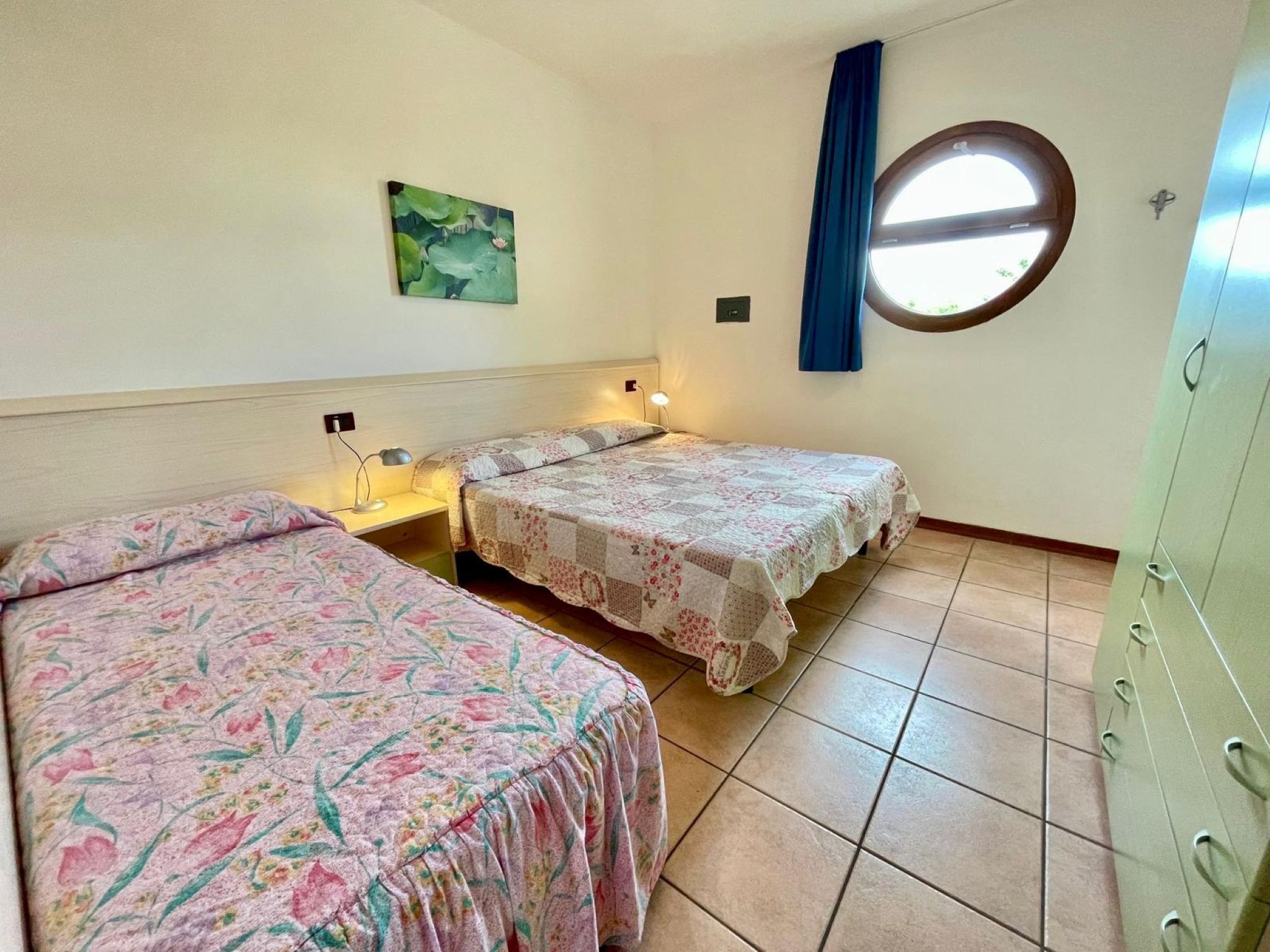 A bedroom with two beds, a nightstand, a wall-mounted lamp, a window with blue curtains, and a wall-mounted circular window.