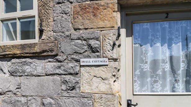 A stone cottage with a sign reading "WELL COTTAGE" and a wooden bench in front. - Well Cottage - 2 bedroom (sleeps 4) (Ashbourne)