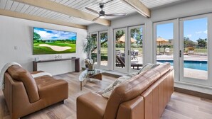 A modern living room with a large flat-screen TV, leather sofas, a glass coffee table, and a ceiling fan. - 6BR Luxury Phoenix Home w Pool, Hot Tub, Games (Phoenix)