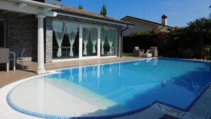 Un área de piscina moderna con una piscina cristalina, un porche cubierto con puertas de cristal y mobiliario exterior.
