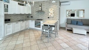 Una cocina moderna con armarios blancos, una isla central y un sofá gris en la zona de estar contigua.