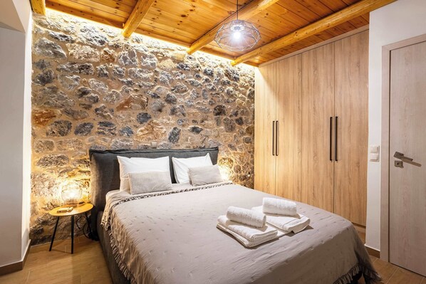 A bedroom with a stone wall, wooden ceiling, a bed with white linens, and a wooden wardrobe.