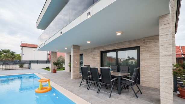 A modern outdoor pool area with a dining table and chairs, surrounded by a stone wall and a glass door. - Luxury villa Marko, family-friendly and pet-friendly modern villa near sea.

 (Vir)