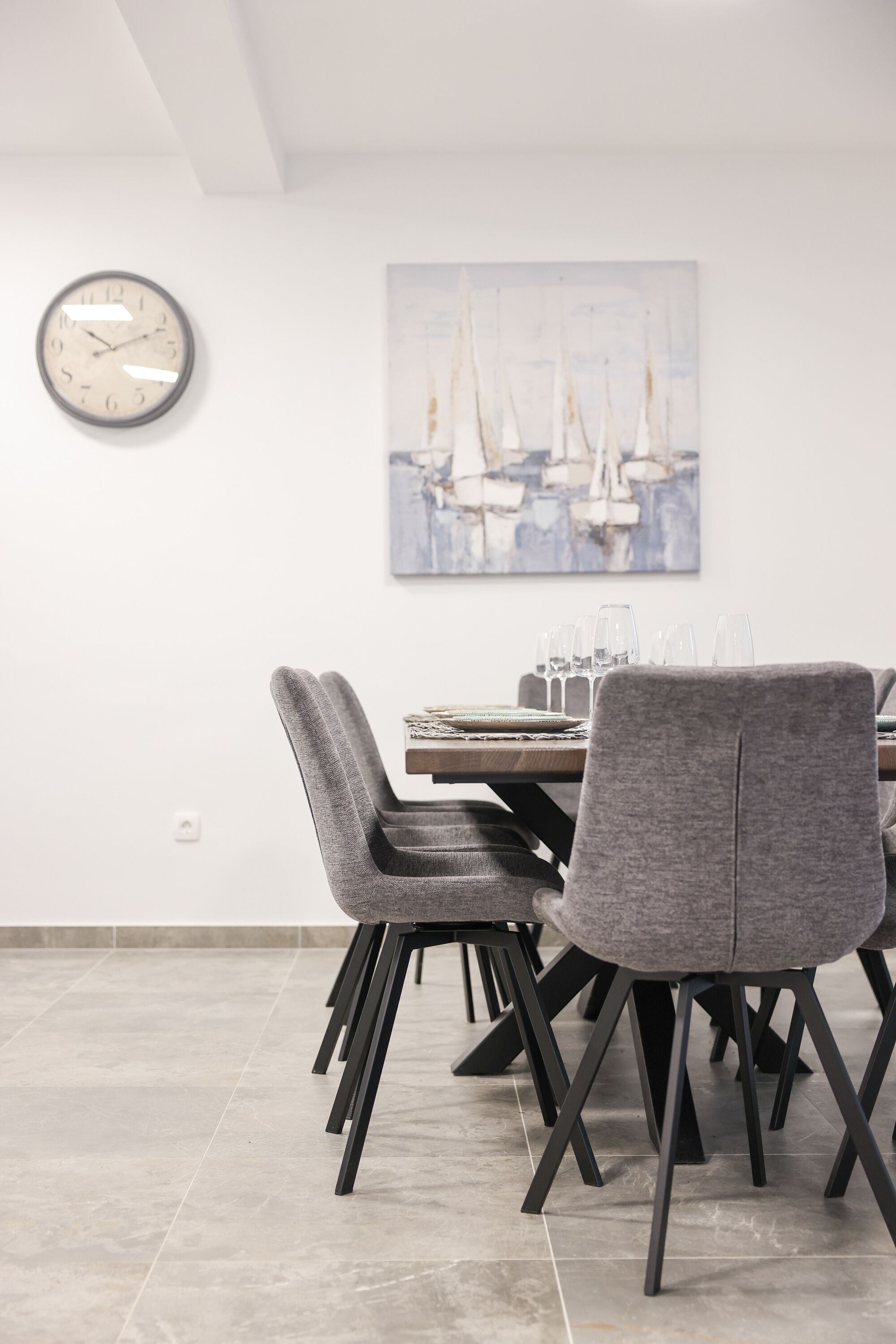 Un coin repas avec une table dressée pour quatre personnes, des chaises à revêtement gris, une horloge murale et un tableau représentant des voiliers.