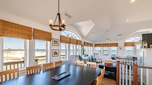 Un'ampia sala da pranzo con un lungo tavolo e sedie, ampie finestre con vista sulla spiaggia e una ventola a soffitto.