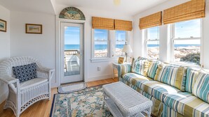 Una stanza con una sedia in vimini, un divano a righe, una ventola a soffitto e vista sulla spiaggia attraverso ampie finestre con tende in bambù.