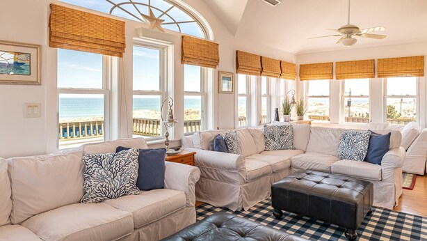 Una stanza luminosa con un'ampia finestra ad arco che si affaccia sulla spiaggia, dotata di ventilatore a soffitto, tappeto a scacchi e diversi divani con cuscini decorati.