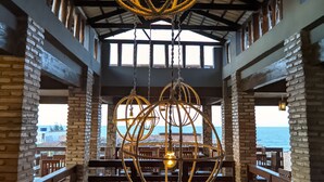 Un restaurant avec un plafond haut, des poutres apparentes et des murs en briques. On y trouve des luminaires sphériques suspendus et une vue sur l’océan grâce à de grandes fenêtres.