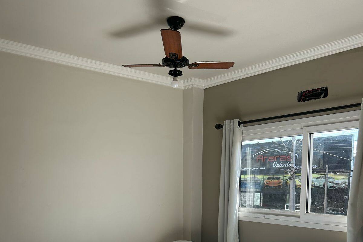 A room with two single beds, a ceiling fan, a window with curtains, and a view of a car dealership outside.