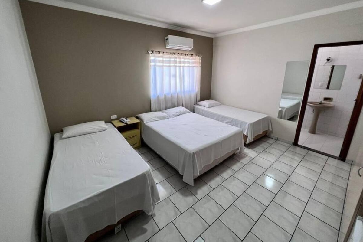 A hotel room with three single beds, a window with curtains, an air conditioning unit, a sink, and a mirror.
