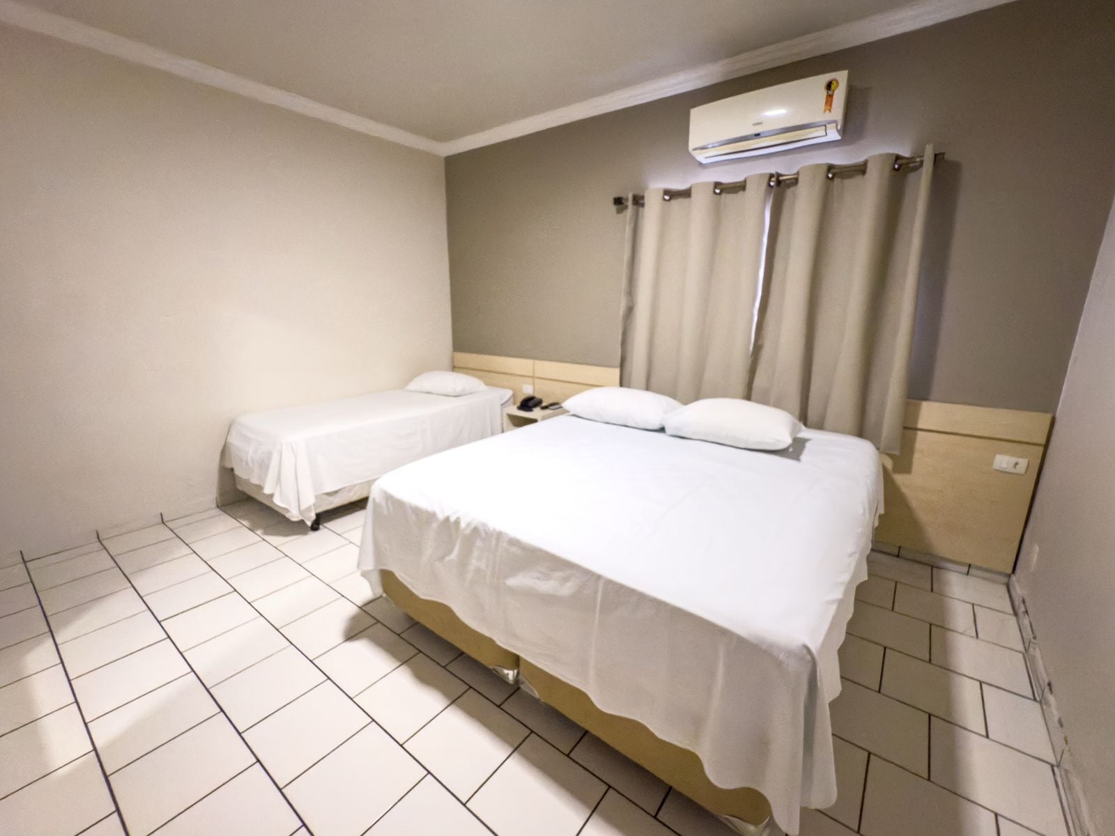 A hotel room with two beds, a white tiled floor, a beige wall, and an air conditioning unit.