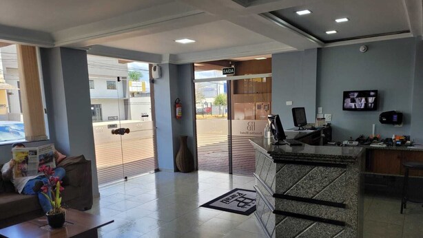 A hotel lobby with a reception desk, a seating area with a coffee table and a potted plant, and a large window providing a view of the street. - Hotel Parati Arapongas (Arapongas)