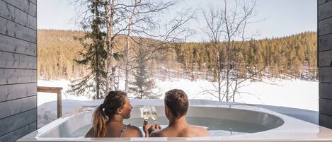A couple in a hot tub with a snowy forest view.