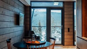 A dining area with a round table and chairs, a television mounted on the wall, and a view of a snowy landscape through a large window. - Kurula's Resort (Pyhätunturi)